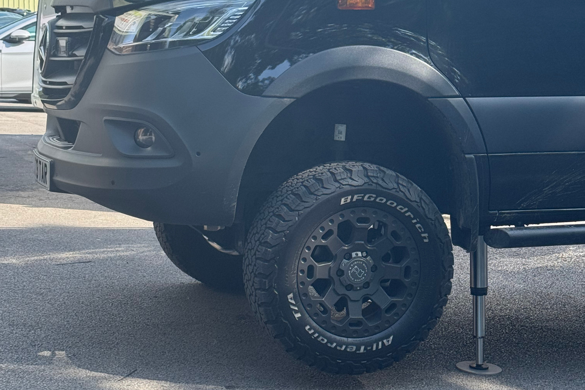 Close-up of a dark gray vehicle with a black BFGoodrich All Terrain T/A tire mounted on a black multi-spoke wheel, parked outdoors on asphalt with the vehicle's side support visible.